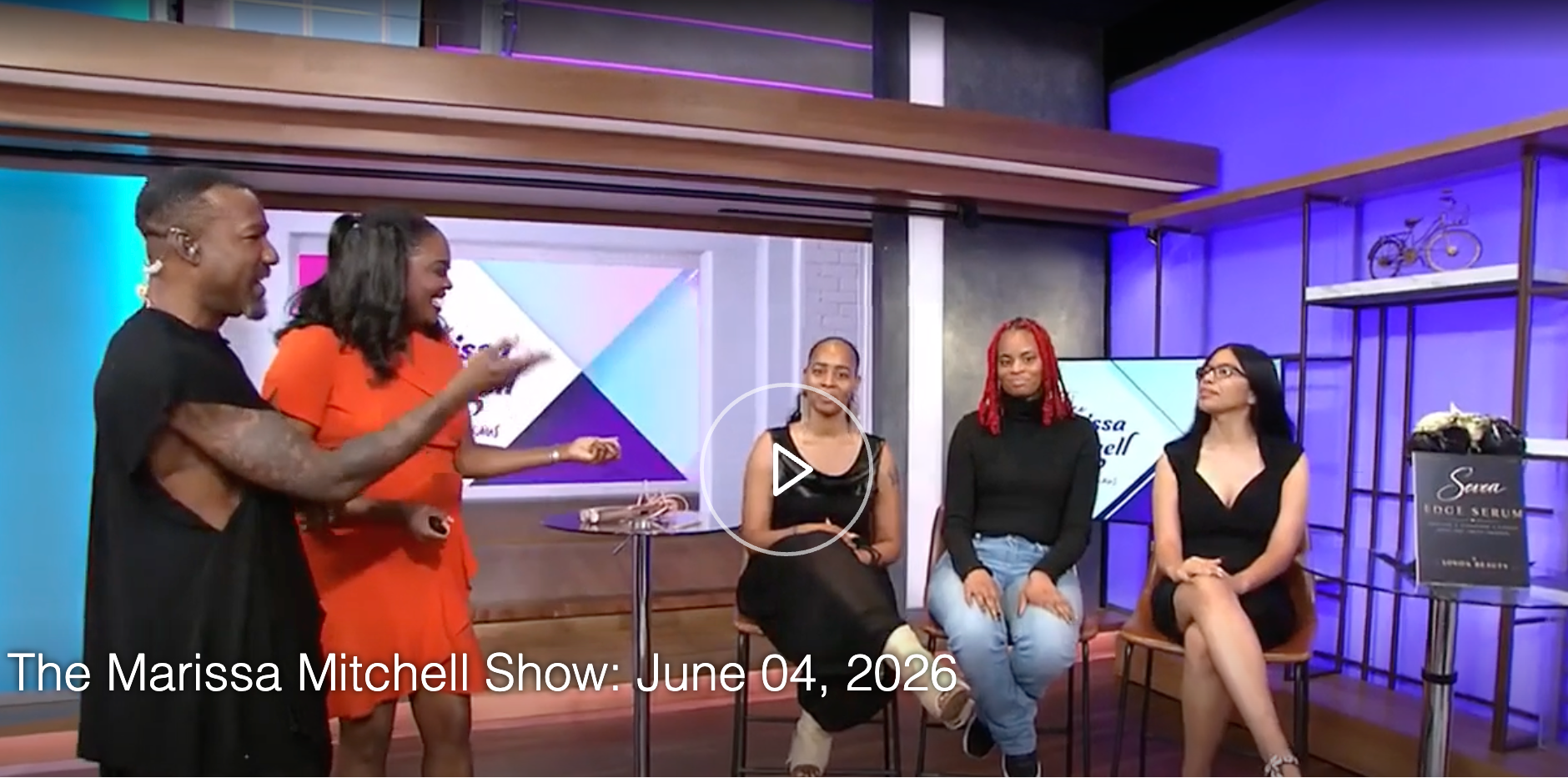 TV show set with four people on a stage, one person standing and three sitting.